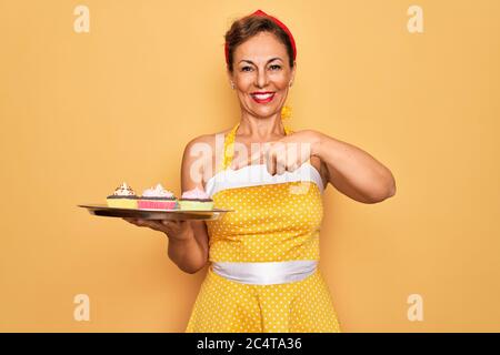 Âge moyen senior épingler une femme portant le style des années 50 rétro tenue plateau avec cupcakes très heureux de pointer avec la main et le doigt Banque D'Images