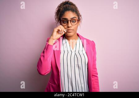 Belle femme d'affaires afro-américaine portant une veste et des lunettes sur fond rose bouche et lèvres fermées comme zip avec les doigts. Secret et silencieux, t Banque D'Images