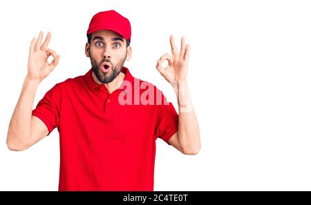 Jeune homme beau avec barbe portant l'uniforme de livraison regarder surpris et choqué faisant ok symbole d'approbation avec les doigts. Expression folle Banque D'Images