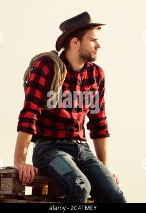 Un agriculteur réfléchi qui pense aux affaires. Ouvrier de Ranch. Éco-exploitation. Concept agricole. Bel homme avec chapeau et tenue de style rustique. Conserver le ranch. La vie au ranch. Cowboy avec lasso corde ciel fond. Banque D'Images