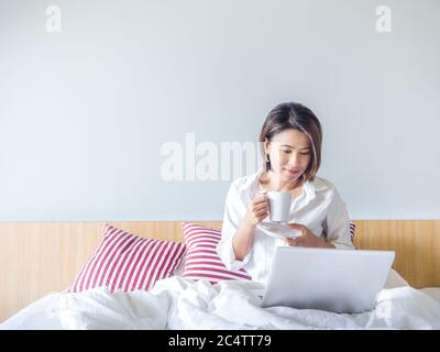Belles femmes asiatiques avec des cheveux courts portant chemise blanche tenant une tasse de café et utilisant ordinateur portable sur un lit en bois dans la chambre blanche dans la maison avec Banque D'Images