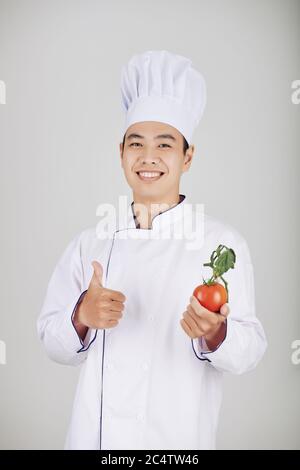 Portrait d'un jeune chef de restaurant positif en uniforme blanc montrant des tomates fraîches et des pouces Banque D'Images