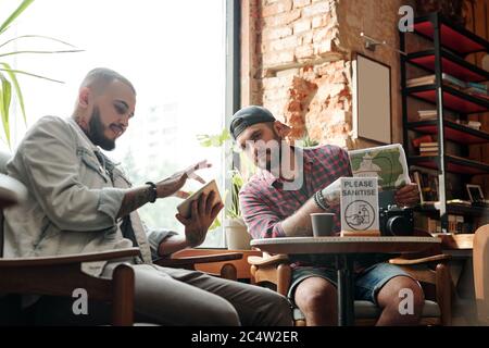Jeune barbu utilisant l'application de voyage sur tablette tout en planifiant un voyage avec un ami dans le café loft Banque D'Images