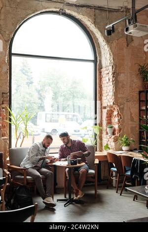 Jeunes amis masculins se reposant dans un café loft et utilisant des appareils modernes tout en discutant des dernières applications Banque D'Images