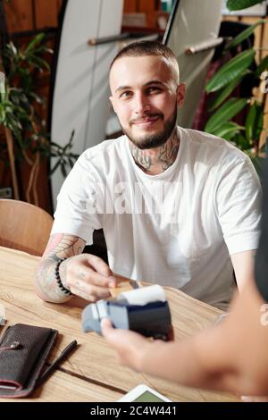 Jeune barbu souriant, en chemise blanche, mettant une carte sans fil au terminal tout en payant le déjeuner au café Banque D'Images