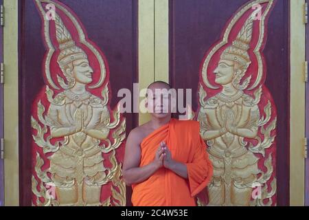 Un moine avec les mains repliées dans une trad. Geste 'wai' thaï à une porte avec deux êtres célestes dans une posture similaire; Wat Mongkon Nimit, ville de Phuket, Thaïlande Banque D'Images