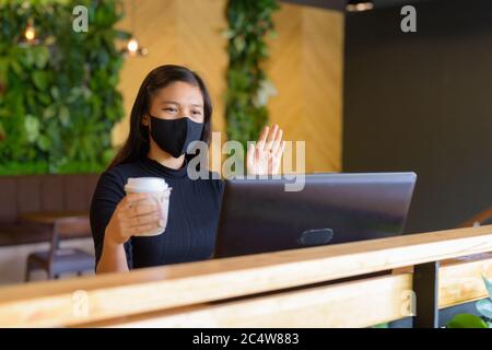 Jeune femme d'affaires asiatique avec appels vidéo masque et assis à distance dans le café Banque D'Images