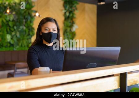 Jeune femme d'affaires asiatique avec masque utilisant un ordinateur portable et assis à distance à l'intérieur du café Banque D'Images