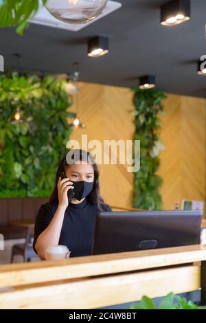 Jeune femme d'affaires asiatique avec masque utilisant un ordinateur portable et parlant au téléphone à l'intérieur du café Banque D'Images