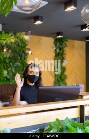 Jeune femme d'affaires asiatique avec appels vidéo masque et assis à distance dans le café Banque D'Images