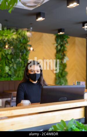 Jeune femme d'affaires asiatique avec masque et ordinateur portable dans le café Banque D'Images