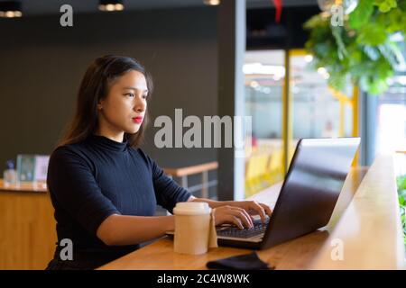 Jeune femme d'affaires asiatique magnifique utilisant un ordinateur portable et assis à distance à l'intérieur du café Banque D'Images