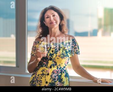 Portrait intérieur d'une belle femme âgée souriante à côté de la fenêtre tenant un verre d'eau Banque D'Images