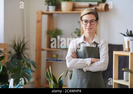Portrait de la jeune femme moderne portant un tablier et regardant l'appareil-photo tout en se tenant debout avec les bras croisés dans la fleur et les plantes magasin, l'espace de copie Banque D'Images