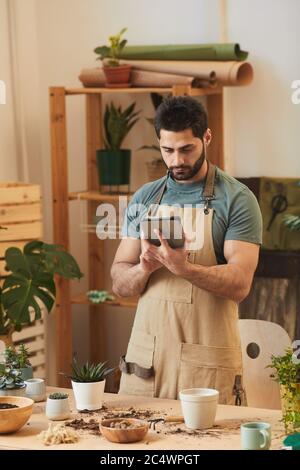 Portrait vertical de l'homme barbu beau portant un tablier et utilisant une tablette numérique tout en se tenant à table avec des plantes de maison et des outils de jardinage Banque D'Images