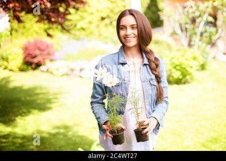 Photo d'une belle jeune femme tenant des fleurs en pot dans ses mains tout en jardinant à la maison. Banque D'Images