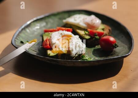 Délicieux petit déjeuner à la maison. Sandwich avec de l'avocat frais tranché au-dessus du pain grillé au seigle avec des tomates cerises et des œufs pochés sur l'assiette verte. Femme Banque D'Images