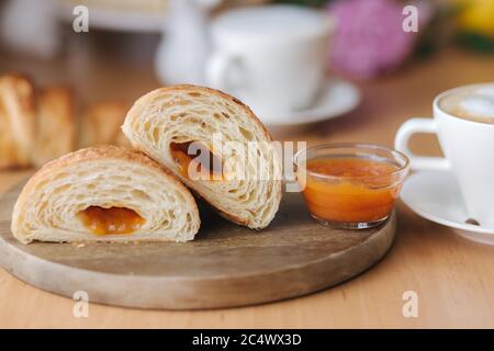 Deux moitiés de croissant fraîchement poêlé sur bois avec confiture d'abricot maison délirante. Fond de cappuccino. Petit déjeuner Banque D'Images