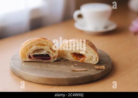 Deux moitiés de croissant fraîchement poêlé sur une planche de bois avec une confiture différente. Fond de cappuccino Banque D'Images
