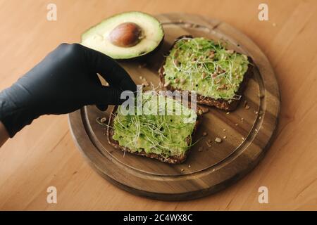 Femme faisant un sandwich vegan avec du pain grillé au seigle avec guacamole et micro verts. Femme saupoudrer de graines de sésame sur le sandwich Banque D'Images