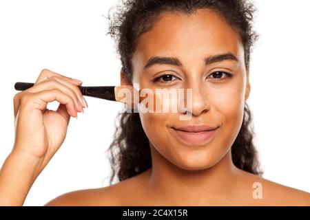 portrait d'une jeune femme à peau foncée appliquant un maquillage liquide avec une brosse sur son visage sur un fond blanc Banque D'Images