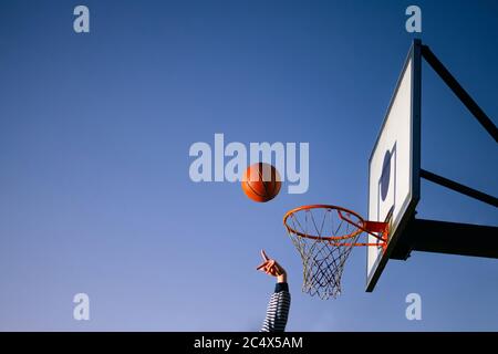 Joueur de balle de basket-ball de rue jetant le ballon dans le panier. Gros plan de la main, boule orange au-dessus du filet de houop avec le ciel bleu en arrière-plan. Concept o Banque D'Images