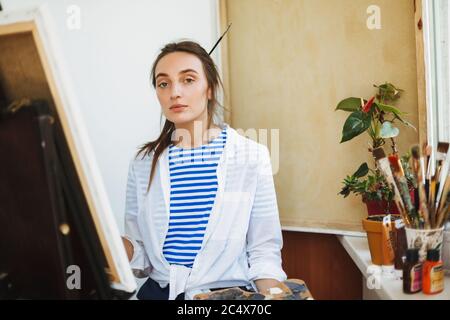 Belle fille en chemise blanche et T-shirt rayé regardant rêveusement dans l'appareil photo tout en dessinant sur le chevalet avec des outils de peinture près sur le seuil de fenêtre à la maison Banque D'Images