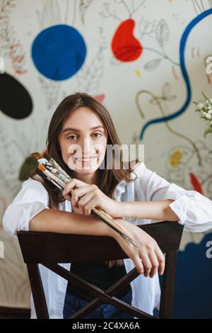 Fille gaie en chemise blanche sur chaise joyeusement regarder dans l'appareil-photo tout en tenant des pinceaux de peinture à la main avec de grands motifs peinture sur le fond à la maison Banque D'Images
