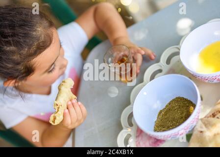 Une petite fille qui boit du thé et mange du pain plat avec de l'huile d'olive et du zaatar à la maison. Petit enfant appréciez la nourriture et les boissons et amusez-vous à l'extérieur dans l'arrière-cour par beau temps. Banque D'Images
