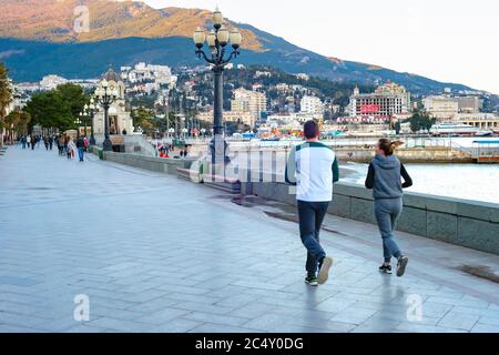 YALTA, CRIMÉE - 3 AVRIL 2018 : couple de jogging sur la promenade de la mer, touristes à pied, église et coucher de soleil paysage urbain par montagne en arrière-plan Banque D'Images
