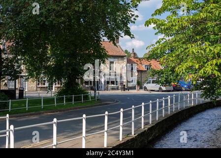 Scène de village avec une rangée de bâtiments en pierre avec des boutiques et un grand arbre en face. Au premier plan, un ruisseau passe par des rails en métal blanc. Banque D'Images