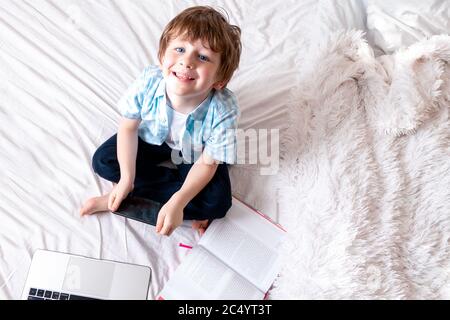 Enseignement en ligne à distance. Un garçon de race blanche heureux avec un livre et un smartphone étudiant et faisant des devoirs à l'école. Assis sur le lit avec un garde-bébé Banque D'Images