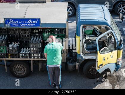 Un homme de traite qui effectue une livraison prend des bouteilles de lait de son véhicule électrique et qui le fournit directement à la porte du client. Une entreprise qui meurt. Banque D'Images