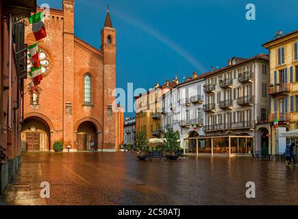 Place de ville pavée humide, maisons anciennes et cathédrale San Lorenzo éclairée par la lumière du soleil sous ciel pluvieux avec arc-en-ciel à Alba, Piémont, Italie du Nord. Banque D'Images