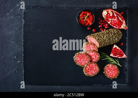 salami enrobé de grains de poivre vert et coupé en tranches sur un panneau de pierre noire et servi avec de la grenade et de la salsa, plat, gros plan, horizonta Banque D'Images