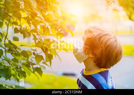 Mignon garçon d'enfant dans le masque médical dans les lilas fleurs nouant à l'odeur des fleurs. Joyeux enfant caucasien à la journée ensoleillée à l'extérieur dans le parc. Fin de la quarantaine Banque D'Images