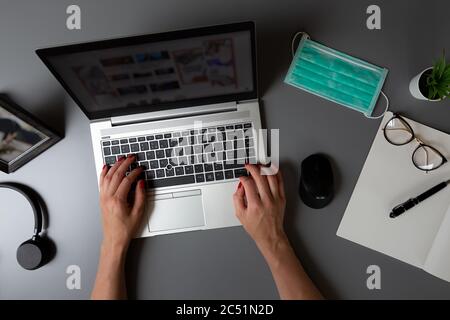 Vue de dessus du bureau à domicile. Les mains des femmes tapant sur un ordinateur portable sur une table grise avec masque de protection du visage. Banque D'Images