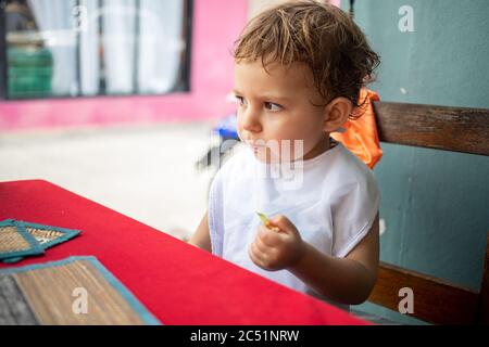 un petit bébé mignon dans un bavoir pensé tout en essayant la nourriture assis à une table sur une terrasse de café de rue Banque D'Images