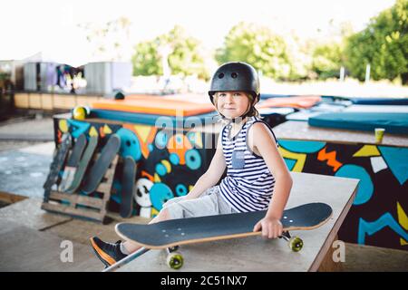 Garçon assis à Skate Park, regardant la caméra. Enfant se reposant avec un skateboard au parc de skate. Enfant assis sur une rampe après l'entraînement sur un skateboard. Tendance Banque D'Images