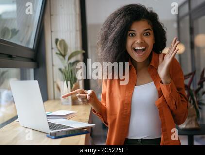 Une femme afro-américaine excitée pointant du doigt sur l'écran d'un ordinateur portable. Emotional hipster shopping en ligne avec des ventes de rabais, de l'argent de retour Banque D'Images