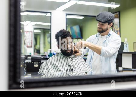 Jeune homme noir heureux en train d'être tondu avec une tondeuse électrique dans un salon de coiffure. Concept de soins de beauté pour hommes. Jeune homme africain souriant, qui devient nouveau Banque D'Images