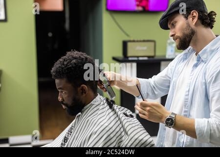 Vue latérale de l'homme avec une coupe de cheveux moderne et élégante, en regardant vers l'avant dans le salon de coiffure. Main de barber gardant le rasoir et coupant les cheveux sur la tête du client. Concept Banque D'Images