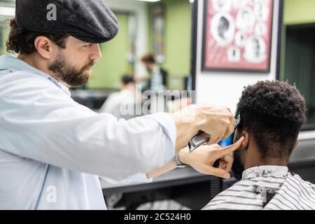 Gros plan du processus de tonte des cheveux dans le salon de coiffure. coiffeur qualifié tenant la tondeuse dans les mains et corrigeant la forme des cheveux à un client masculin assis o Banque D'Images