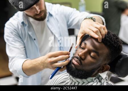 Vue latérale d'un homme sérieux avec une coupe de cheveux moderne et élégante, en regardant vers l'avant dans le salon de coiffure. Main de barber gardant le rasoir droit et en coupant des rayures tendance Banque D'Images