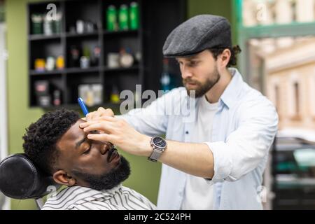 Vue latérale d'un homme sérieux avec une coupe de cheveux moderne et élégante, en regardant vers l'avant dans le salon de coiffure. Main de barber gardant le rasoir droit et en coupant des rayures tendance Banque D'Images