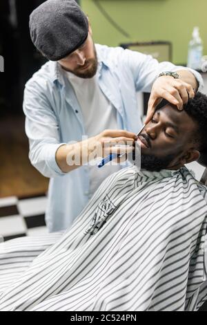 Vue latérale d'un homme sérieux avec une coupe de cheveux moderne et élégante, en regardant vers l'avant dans le salon de coiffure. Main de barber gardant le rasoir droit et en coupant des rayures tendance Banque D'Images