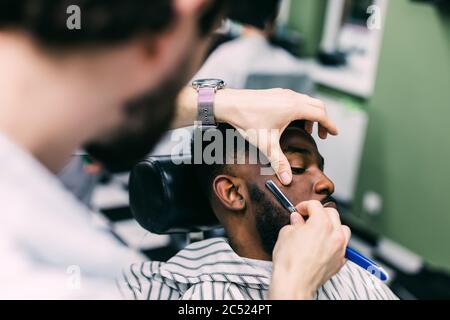 Vue latérale d'un homme sérieux avec une coupe de cheveux moderne et élégante, en regardant vers l'avant dans le salon de coiffure. Main de barber gardant le rasoir droit et en coupant des rayures tendance Banque D'Images