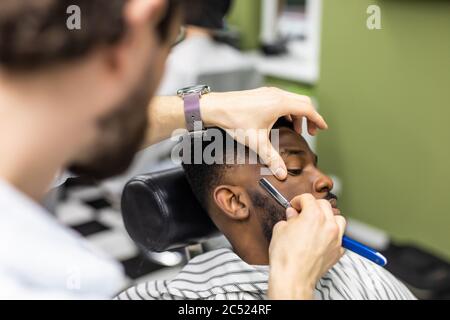 Vue latérale d'un homme sérieux avec une coupe de cheveux moderne et élégante, en regardant vers l'avant dans le salon de coiffure. Main de barber gardant le rasoir droit et en coupant des rayures tendance Banque D'Images