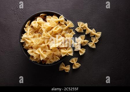 Tas de pâtes farfelle crues qui se répande d'un bol noir sur fond texturé brun. Vue du dessus. Banque D'Images