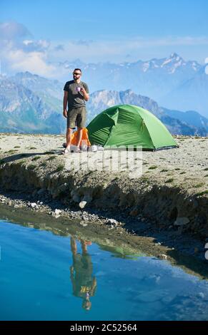 Voyageur tenant une tasse de thé en se tenant à côté de tente touristique, beau lac bleu dans la vallée de montagne. Randonneur masculin dans un short réfléchissant dans l'eau cristalline. Concept de voyage, randonnée, alpinisme et camping. Banque D'Images